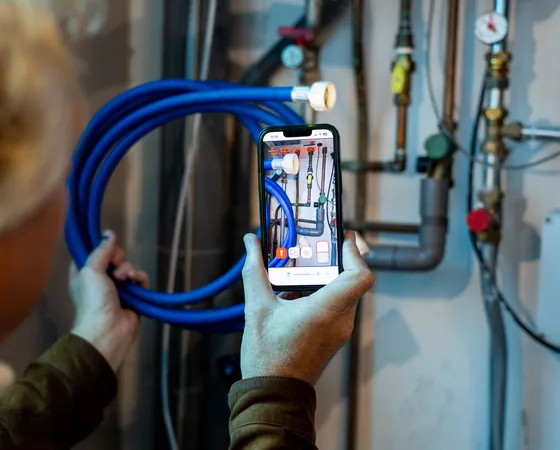 Jemand hält ein Smartphone vor eine technische Anlage mit Leitungen und Messgeräten. Auf dem Bildschirm ist zu sehen, dass ein Foto der Situation aufgenommen wird. Das Bild zeigt einen Moment, in dem jemand eine technische Umgebung dokumentiert.