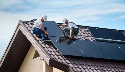 Twee monteurs plaatsen zonnepanelen op het dak van een woonhuis. Ze dragen veiligheidshelmen en werken geconcentreerd aan de montage. Het beeld toont een praktische werksituatie in de buitenlucht, gericht op installatie en verduurzaming van een woning.