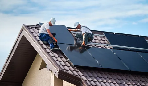 Zwei Monteure installieren Solarmodule auf dem Dach eines Wohnhauses. Sie tragen Schutzhelme und arbeiten konzentriert an der Montage. Das Bild zeigt eine praktische Arbeitssituation im Außenbereich, die auf Installation und die nachhaltige Modernisierung