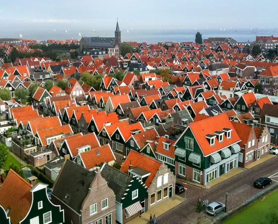 Ein niederländisches Dorf mit traditionellen Häusern mit roten Dachziegeln und grünen Fassaden. Schmale Straßen und Grachten verlaufen durch das Viertel. Im Hintergrund steht eine Kirche mit Turm, außerdem ist Wasser mit Segelbooten in der Ferne zu sehen.
