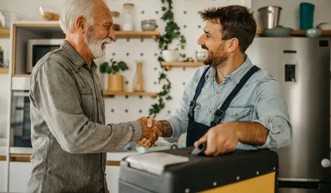 Ein Monteur und ein Bewohner schütteln sich in einer wohnlichen Küche die Hand. Der Monteur hat seinen Werkzeugkoffer dabei. Das Bild vermittelt Vertrauen, persönlichen Kontakt und eine abgeschlossene Service- oder Wartungssituation.