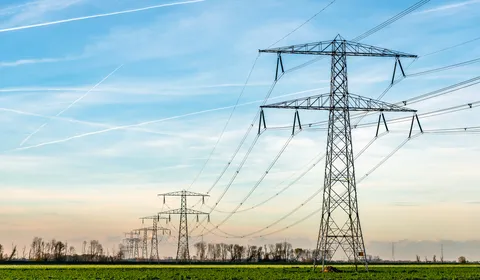 Een rij hoogspanningsmasten met elektrische kabels strekt zich uit over een groen veld. De lucht is blauw met enkele wolken en vliegtuigstrepen. Op de achtergrond staan bomen en een lichte gloed van de ondergaande zon.