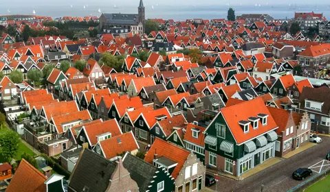 Een Nederlands dorp met traditionele huizen met rode dakpannen en groene gevels. De smalle straten en grachten lopen door de wijk. Op de achtergrond staat een kerk met een toren en is water zichtbaar met zeilboten in de verte.